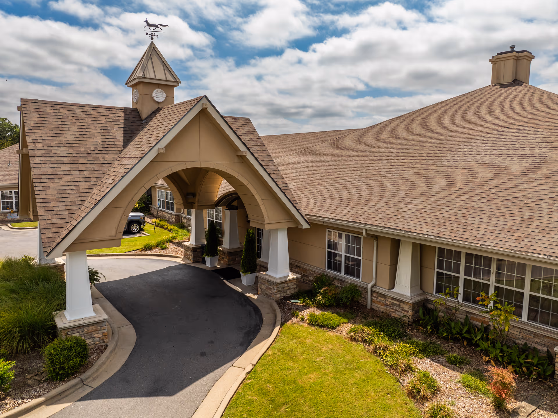 Covered entrance/porte-cochere of a single-story senior living building with a driveway and landscaped lawn under a partly cloudy sky.