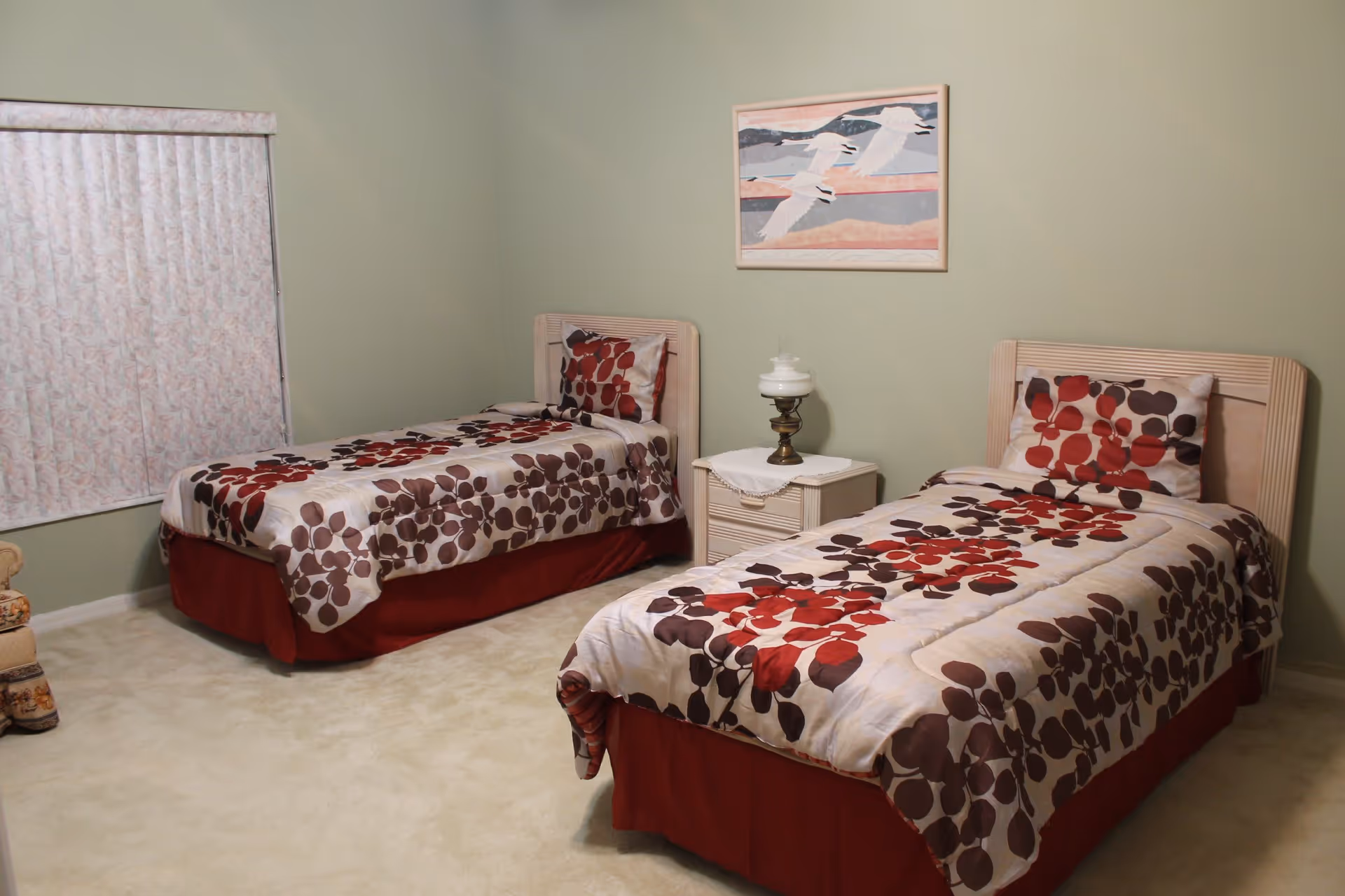 A bedroom with two twin beds in matching floral bedspreads, a nightstand with a lamp between them, and a seagull painting on a pale green wall.