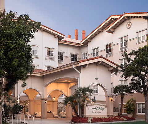 Exterior view of Belmont Village Senior Living Burbank building with arched entrance, palm trees, and well-maintained landscaping under a clear sky.