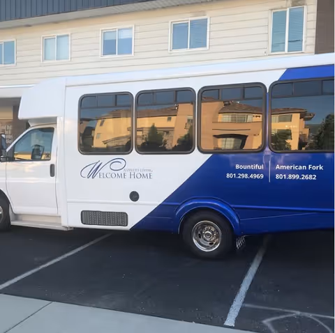 A white-and-blue Welcome Home Assisted Living shuttle van parked outside a light-colored apartment building.