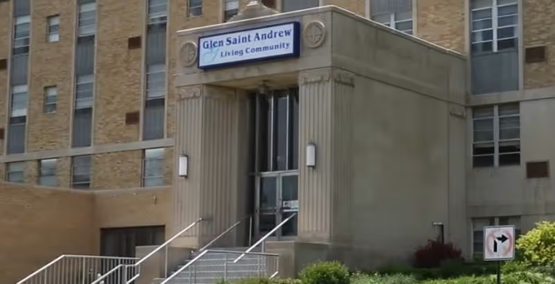 Entrance of a multi-story brick building with a sign above the door that reads 'Glen Saint Andrew Living Community'. There are stairs leading up to the entrance with metal railings on both sides, and some greenery in front of the building.