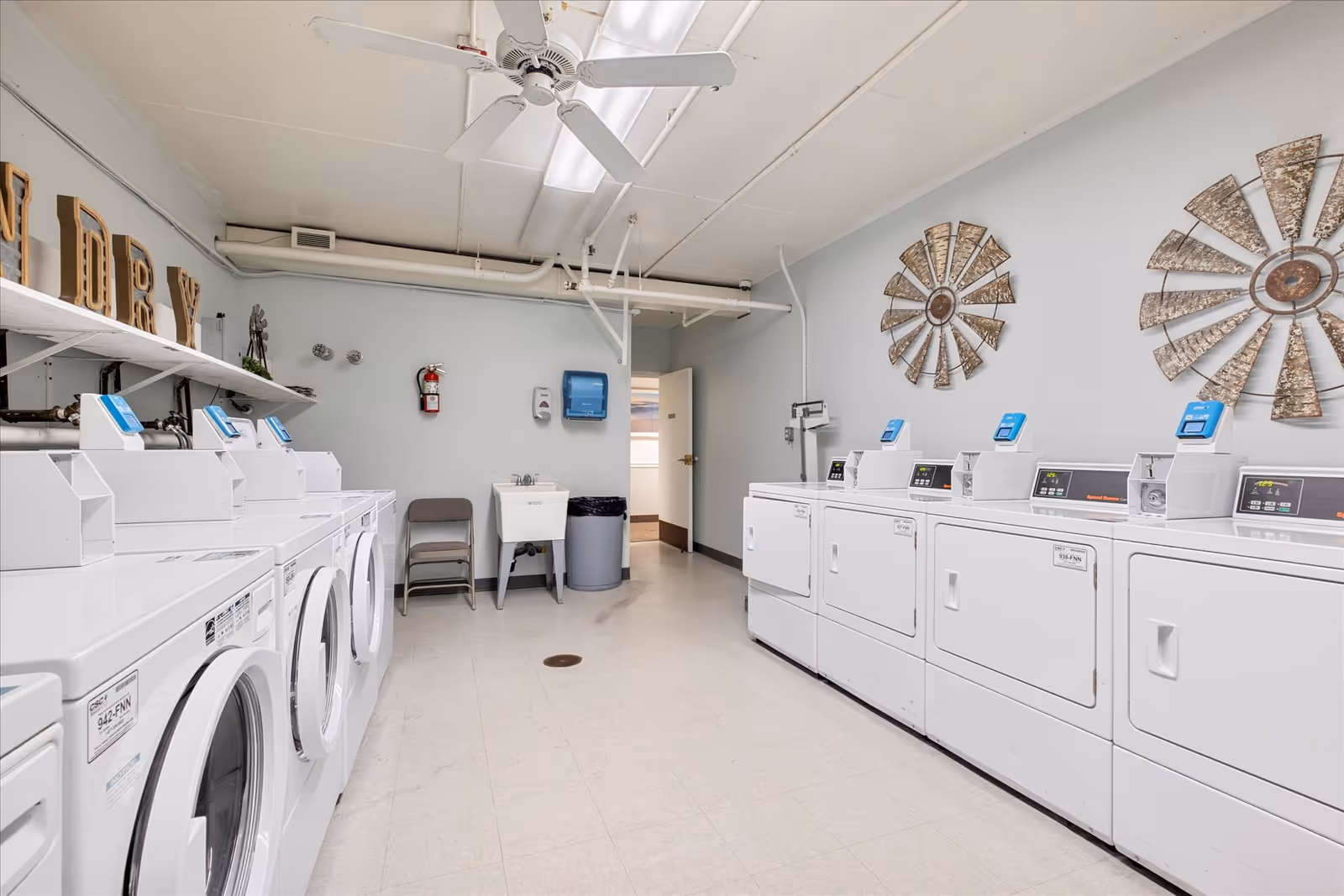Laundry room with multiple white washing machines and dryers lined up on both sides of the room. There is a ceiling fan, a utility sink, a trash can, a folding chair, and decorative metal wall art resembling windmill blades on the walls.