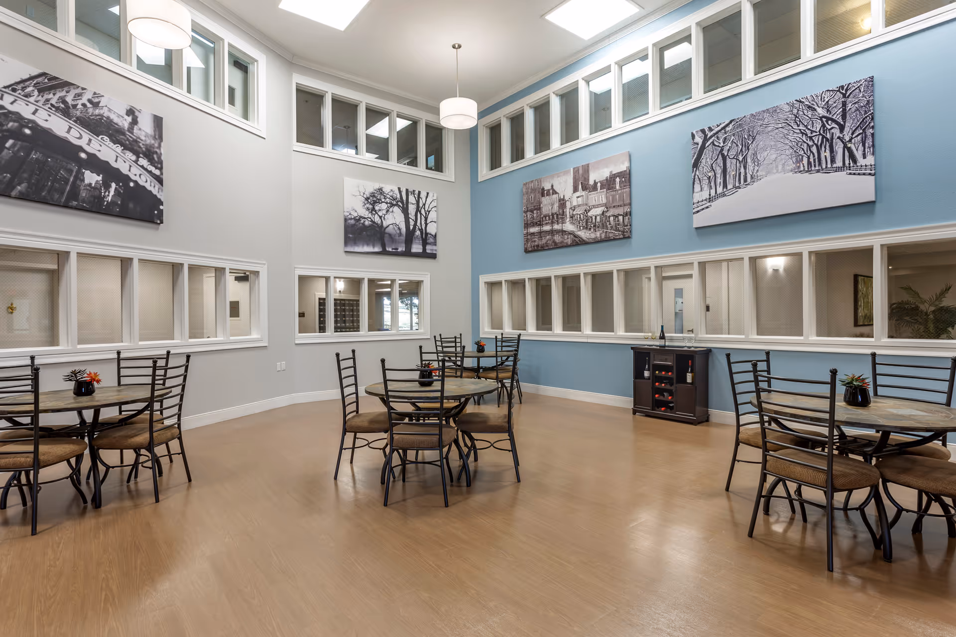 Bright communal dining room with round tables and metal chairs, high windows, a blue accent wall and black-and-white artwork.