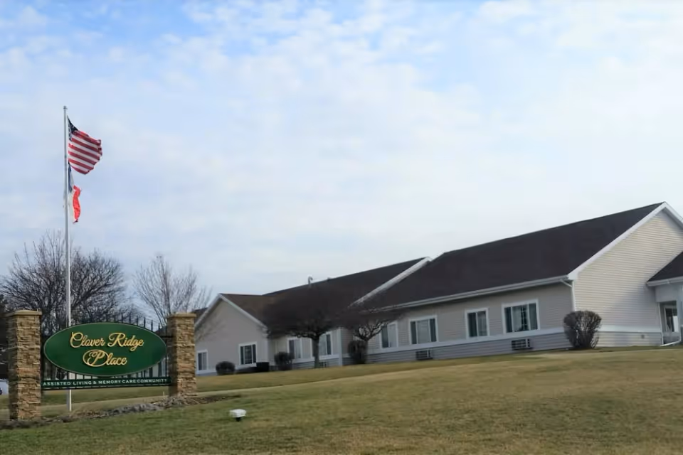Front exterior of Clover Ridge Place senior living building with a lawn, flagpoles, and a green entrance sign.