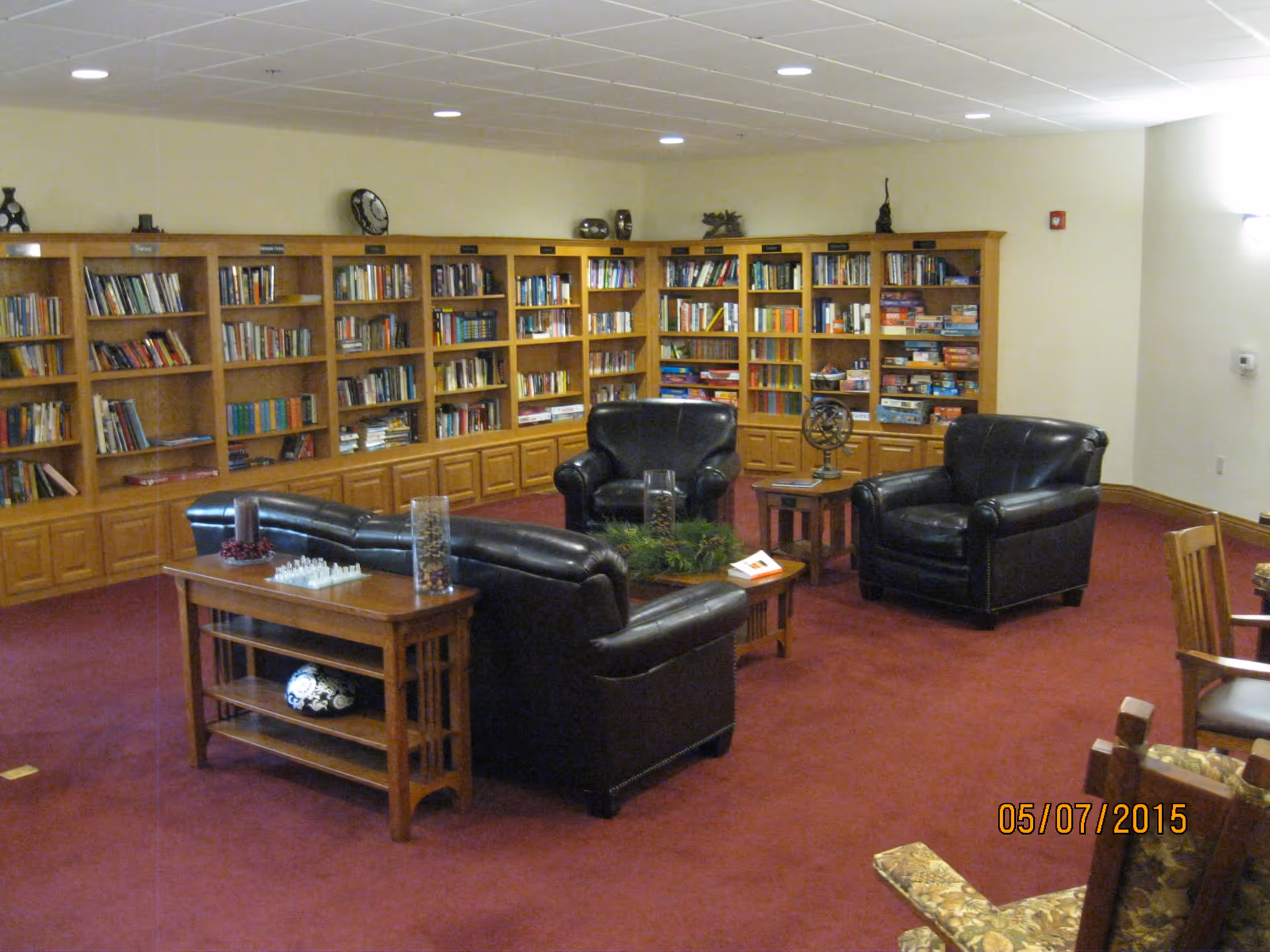 A cozy library or reading room with wooden bookshelves filled with books and board games along the back wall. The room has a red carpet and several black leather armchairs and a sofa arranged around wooden tables. Decorative items and a chess set are placed on the tables. The walls are light-colored with ceiling lights providing illumination.