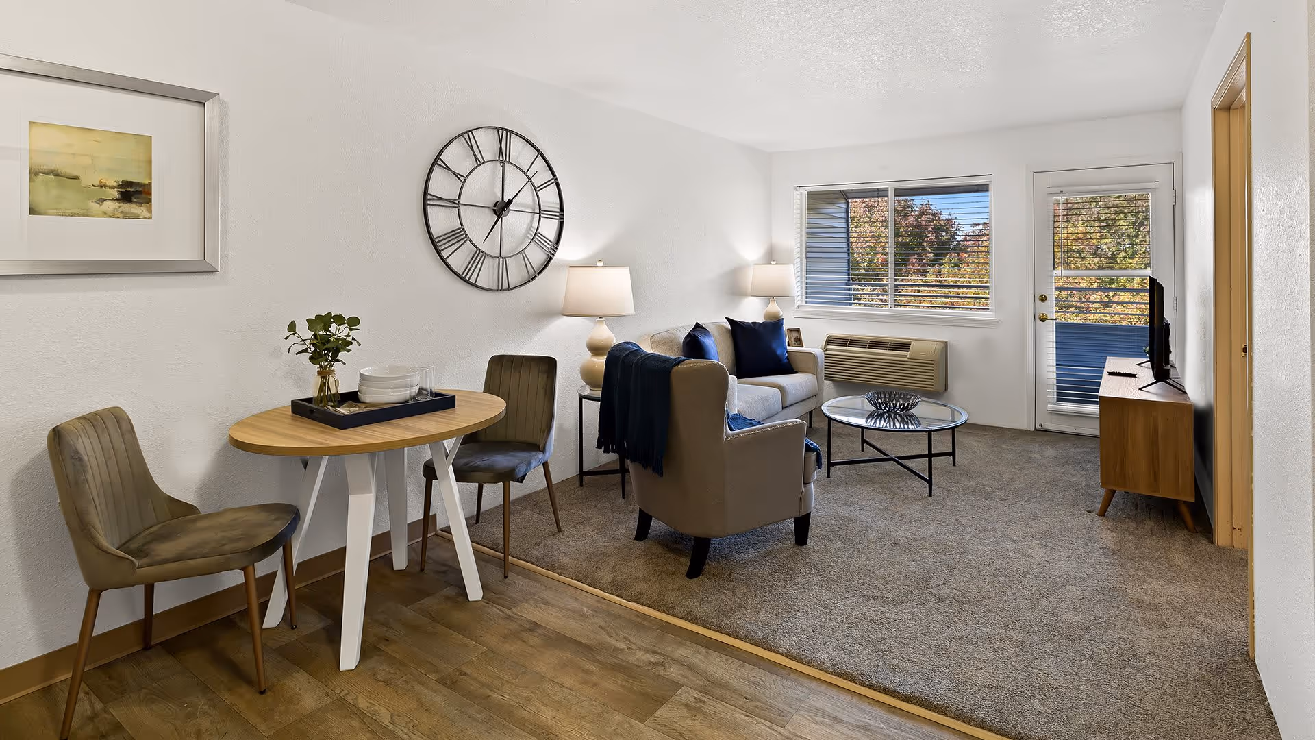 A cozy living room and dining area with a small round wooden table and two chairs on a wooden floor. The living room has a beige armchair, a beige sofa with blue cushions, a glass coffee table, two lamps, a wall clock, and a TV on a wooden stand. A window and a glass door with blinds show trees outside.