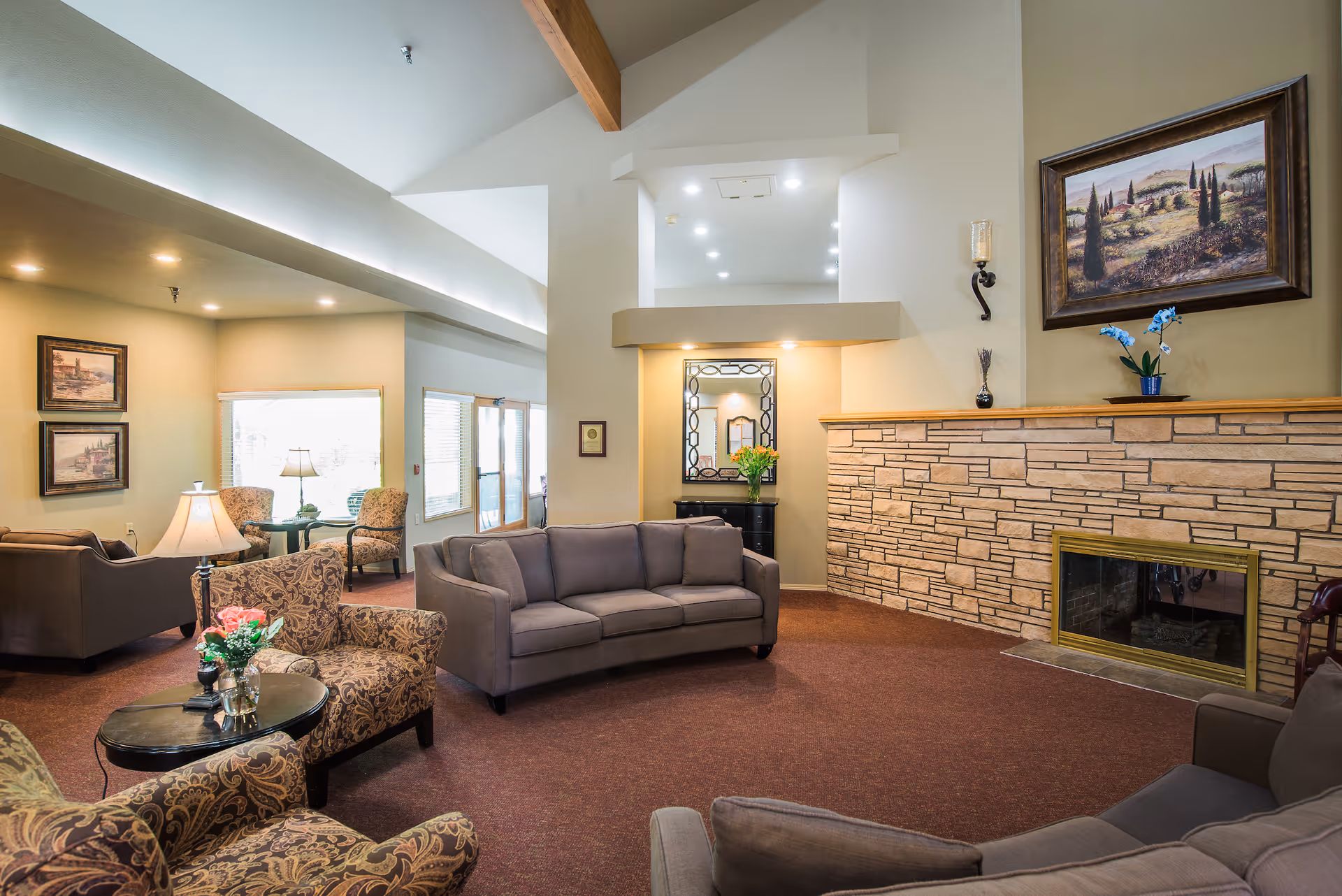 A spacious living room area with multiple seating options including a gray sofa, patterned armchairs, and a small round table with a lamp and flowers. The room features a stone fireplace with a framed landscape painting above it, beige walls, and a high ceiling with exposed wooden beams. There is a mirror and a small black cabinet with flowers on it in a recessed wall area, and large windows letting in natural light.