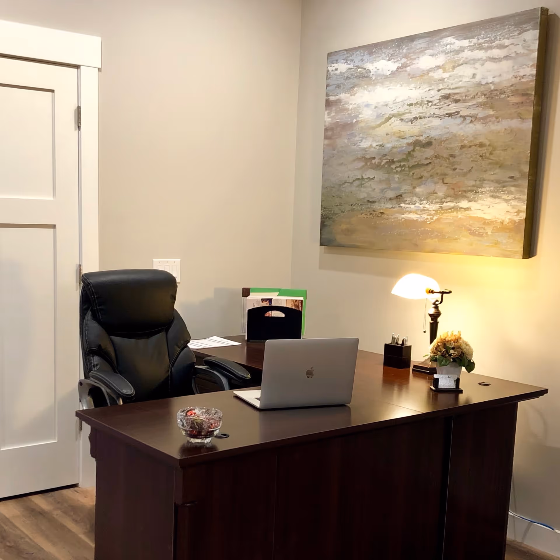 A small office space with a dark wooden desk, a black leather office chair, a silver laptop, a glass bowl with candies, a small lamp, a flower arrangement, and a large abstract painting on the wall. There is a closed white door to the left.
