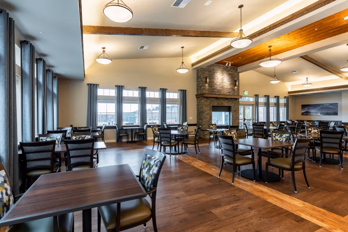 Spacious dining room with multiple tables and chairs, a central stone fireplace, large windows and pendant lighting.