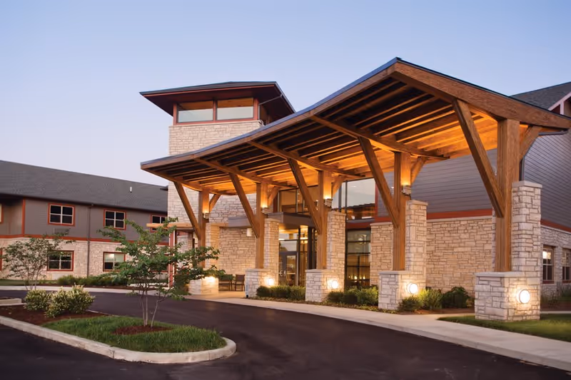 Exterior view of Wellbrooke of Westfield senior living facility at dusk, featuring a large covered entrance with wooden beams and stone pillars, illuminated by warm lights, with landscaped greenery and a paved driveway.