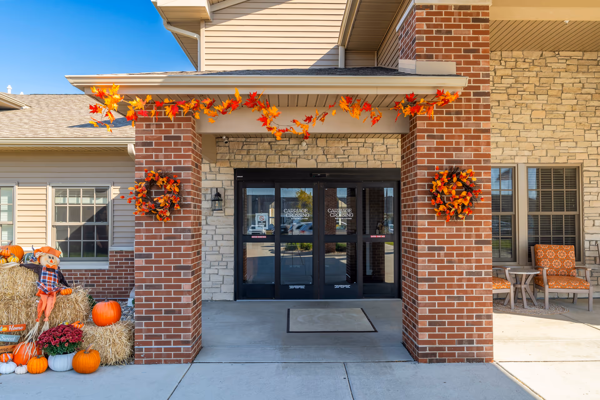 Entrance to The Carriages of Rochester facility decorated with autumn-themed wreaths and garlands. There are pumpkins, hay bales, and a scarecrow on the left side, and two orange cushioned chairs with a small table on the right side under a covered porch.