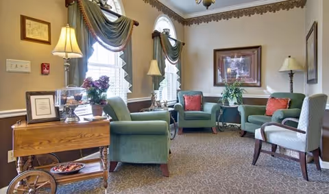 A cozy senior living facility lounge with green upholstered armchairs and a patterned carpet. The room features two arched windows with green drapes, two table lamps on side tables, framed artwork on the walls, and a wooden cart with decorative items including a potted plant and a glass container.