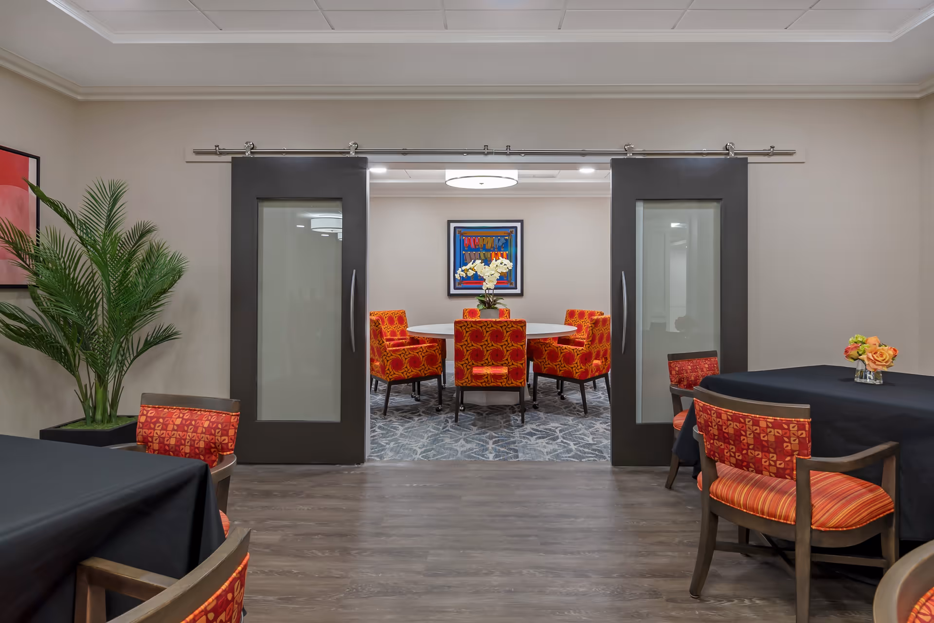 Interior view of a senior living facility dining area with a round table surrounded by six red and orange patterned chairs in a carpeted room visible through two large sliding glass doors. In the foreground, there are tables covered with black tablecloths and chairs with red and orange cushions. A potted plant and framed artwork are on the walls.