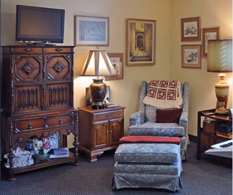 Cozy living-room corner with an upholstered armchair and ottoman, wooden cabinets and side tables, table lamps, and framed artwork on the walls.