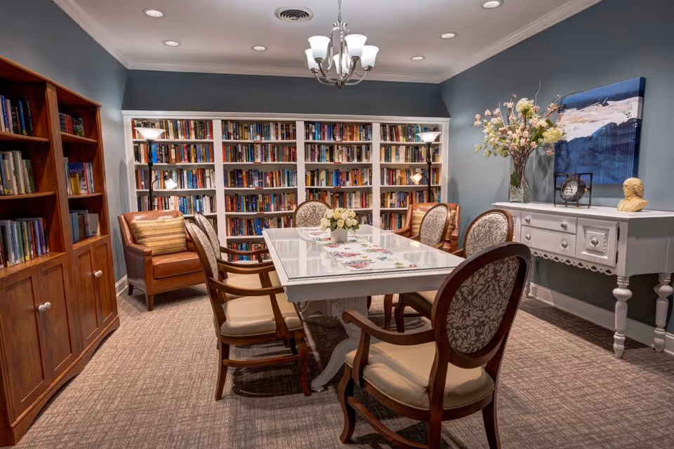 A cozy library room with a white table surrounded by six upholstered chairs. The walls are painted blue-gray and lined with white bookshelves filled with books. There is a wooden cabinet on the left, a brown leather armchair with a pillow, two standing lamps, and a white console table on the right decorated with a vase of flowers, a clock, and a bust sculpture. A chandelier hangs from the ceiling.