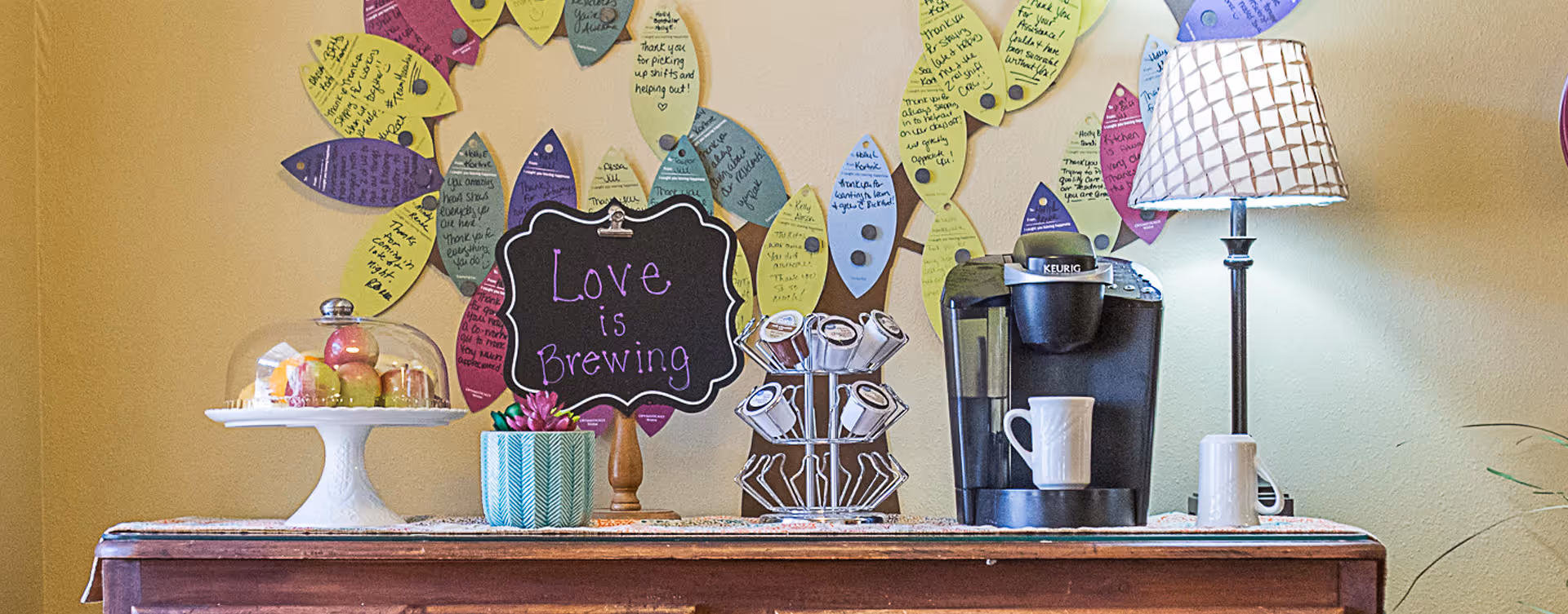 A coffee station on a wooden table featuring a Keurig coffee maker with a white mug, a lamp with a patterned shade, a small potted plant, a cake stand with a glass cover holding apples, and a decorative chalkboard sign that reads 'Love is Brewing'. Colorful paper leaves with handwritten notes are pinned on the wall behind the table.