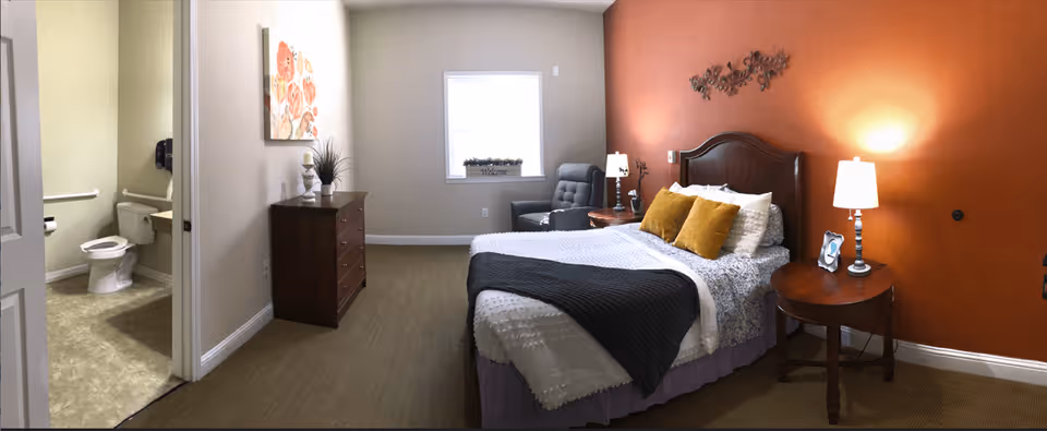 A cozy bedroom with a bed featuring white and patterned bedding, two yellow pillows, and a dark throw blanket. There are two wooden nightstands with lamps on either side of the bed. A dark armchair is positioned near a window with a small plant on the windowsill. A wooden dresser with decorative items and a floral painting is against the opposite wall. To the left, an open door reveals a bathroom with a toilet and grab bars.