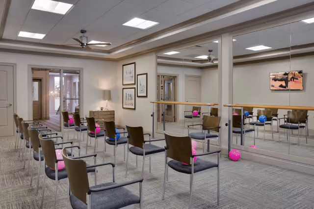A spacious exercise room with rows of chairs arranged facing a mirrored wall with a ballet barre. Colorful exercise balls are placed on some chairs and on the floor. The room has a ceiling fan, framed artwork on the walls, and a door leading to another area.