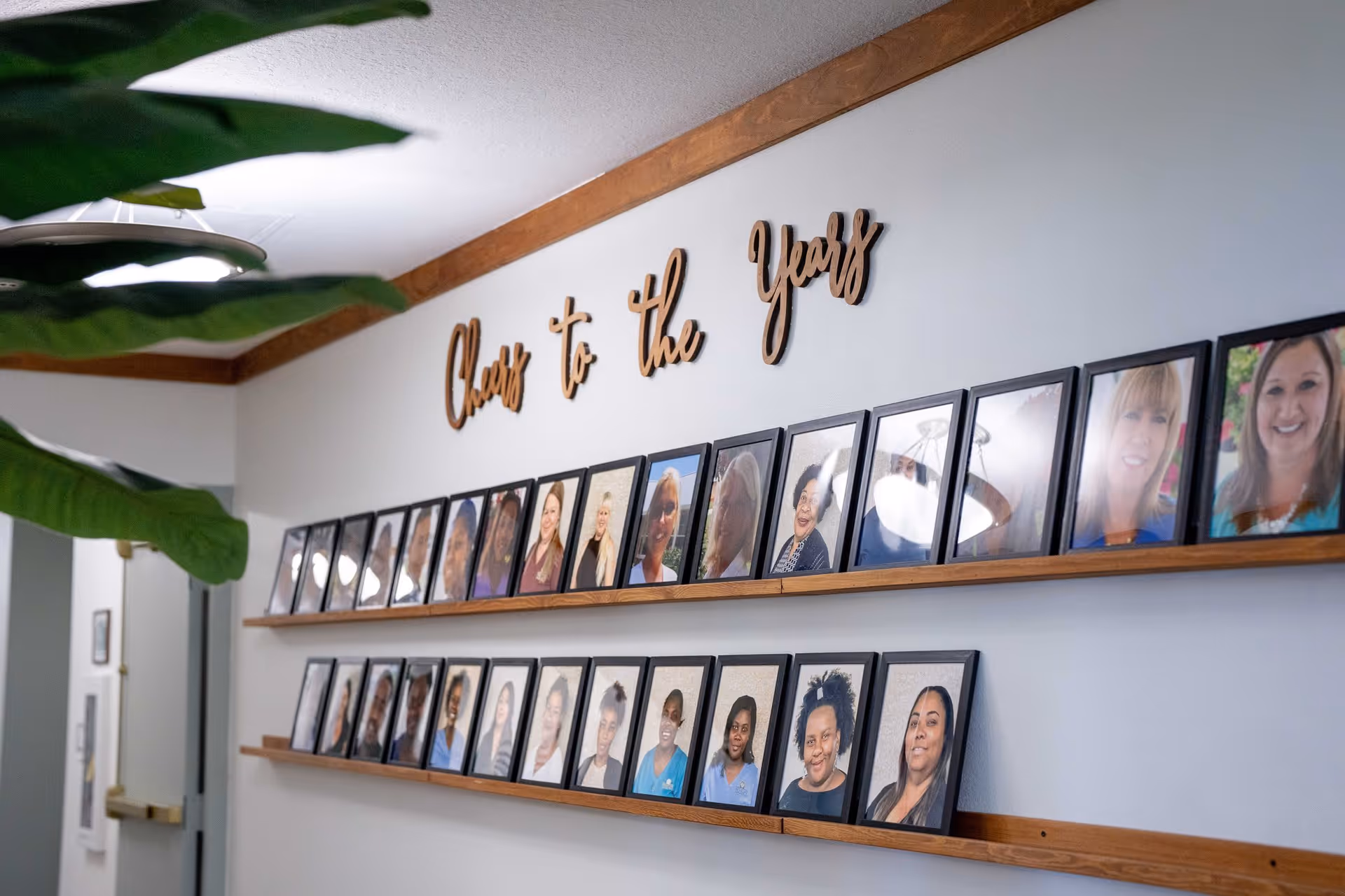 A hallway wall displaying two wooden shelves with framed photographs of various people. Above the shelves, wooden cursive letters spell out 'Cheers to the Years'. A large green plant is partially visible on the left side of the image.