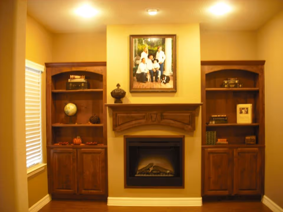 Cozy interior room with a built-in wooden fireplace mantel and two matching wooden bookshelves on either side. The shelves hold decorative items including a globe, books, and small ornaments. Above the fireplace hangs a framed family portrait. The walls are painted a warm beige color and the floor is wooden.