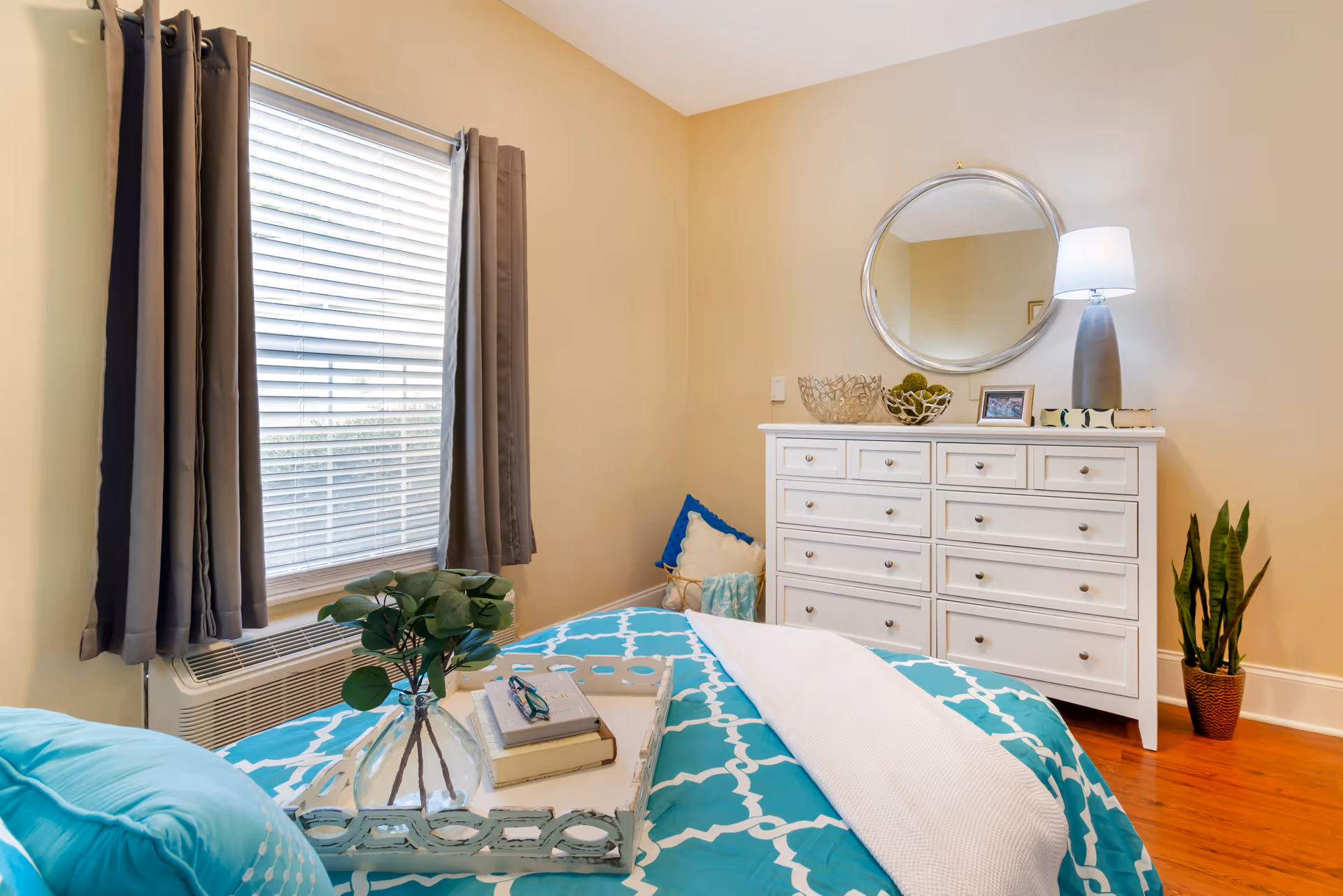 Bright bedroom with a teal patterned bedspread, white dresser topped with a round mirror and lamp, and a window with gray curtains.
