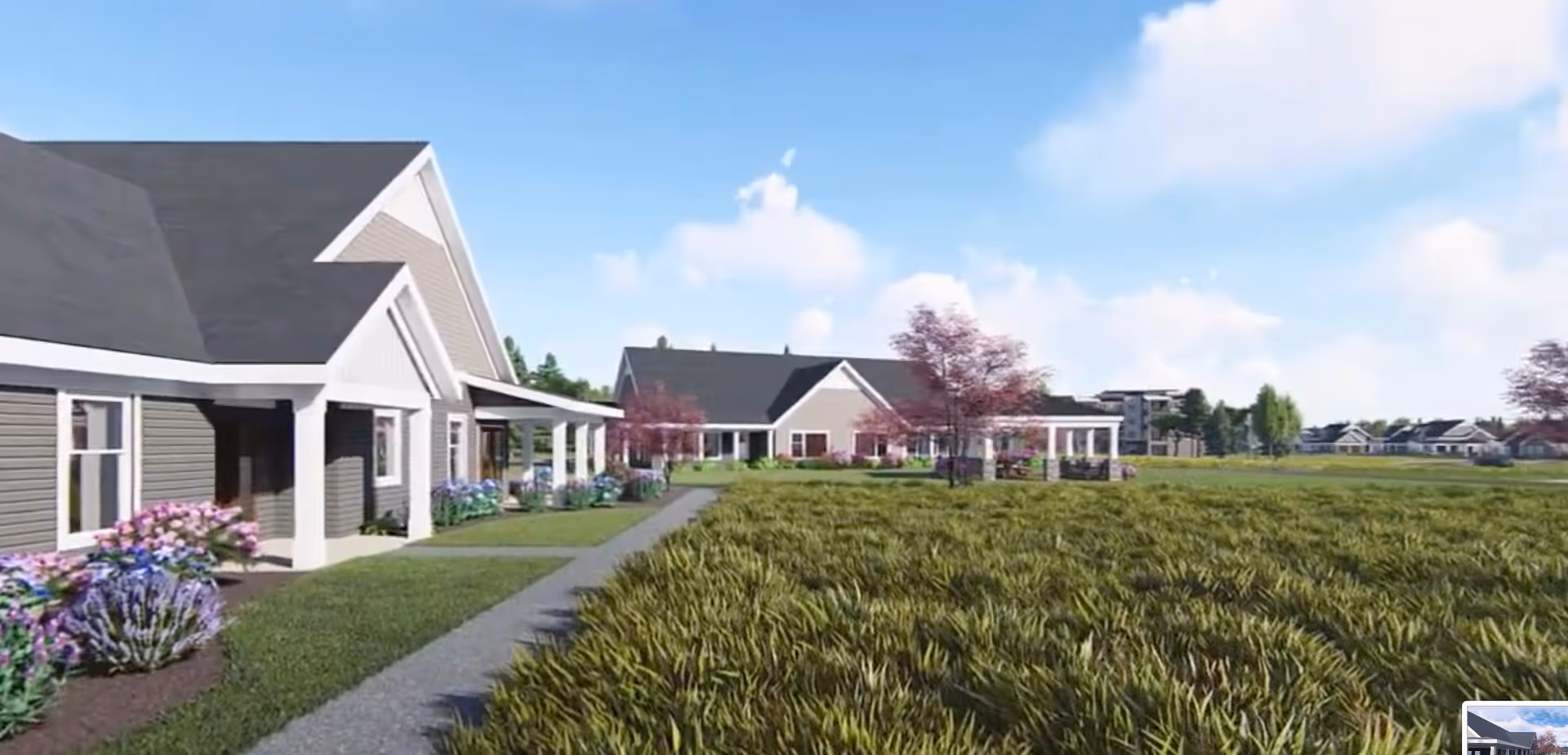 Exterior view of a senior living campus with single-story buildings, a walkway, landscaped flowerbeds, and a large grassy field under a blue sky.