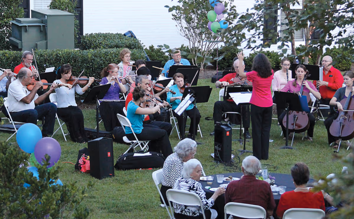 A conductor leads an outdoor orchestra as seated musicians play and residents watch at tables on the lawn.
