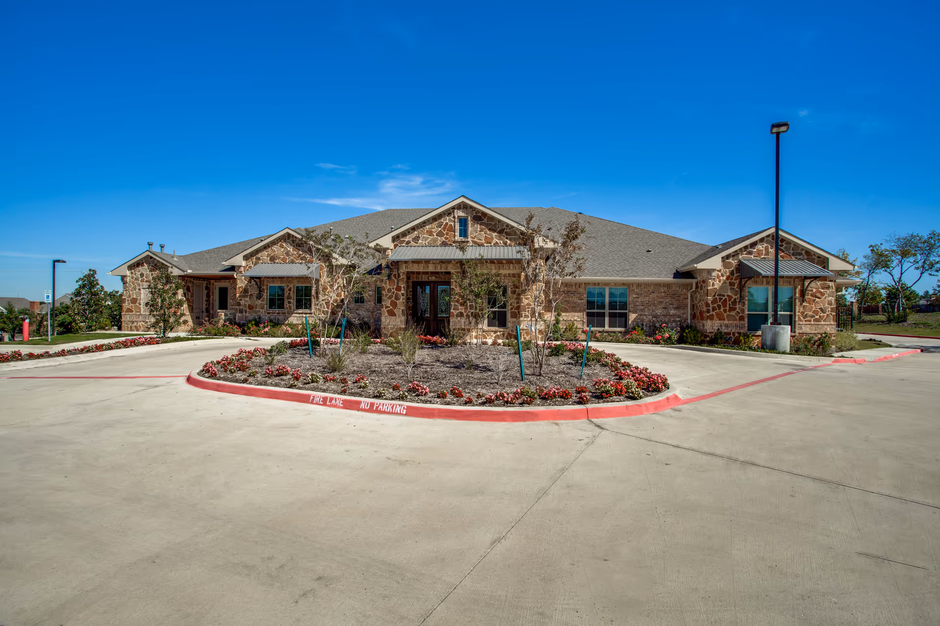 Front exterior view of a single-story stone and brick building with a gray shingled roof, surrounded by a concrete driveway and landscaped with small trees and flower beds under a clear blue sky.