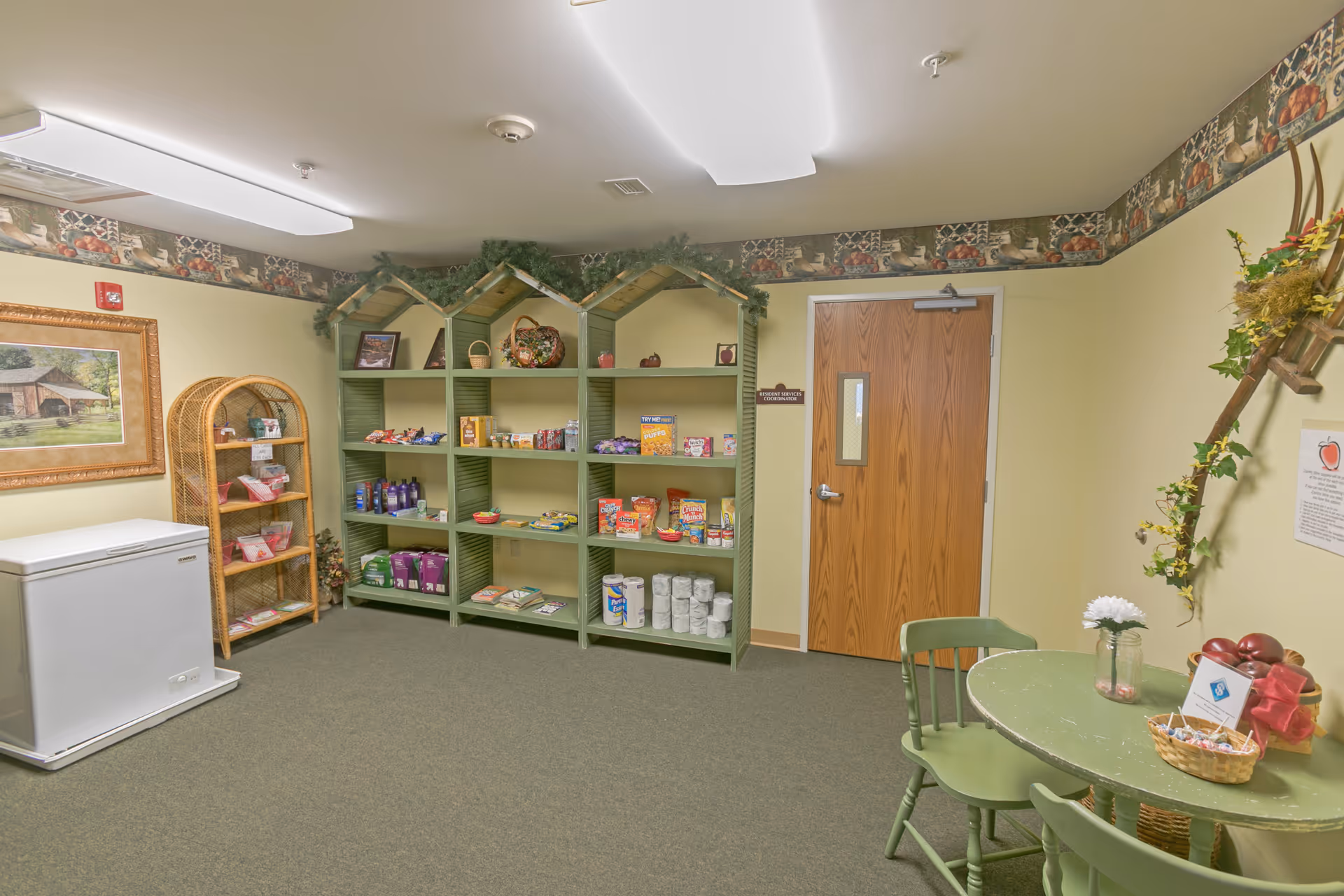A small room with green shelves shaped like houses stocked with various snacks and household items, a wicker shelf with additional items, a white chest freezer, a green round table with three matching chairs, and decorative plants and wall art. The room has beige walls with a decorative border near the ceiling and a wooden door.
