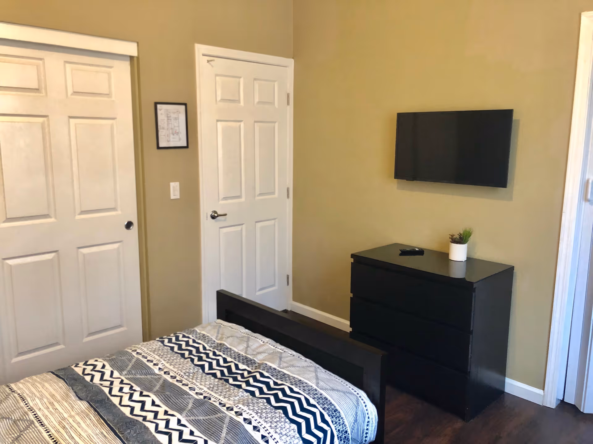 A bedroom with a bed covered in a patterned quilt, a black dresser with a small potted plant and a TV mounted on the beige wall above it. There are two white doors, one sliding closet door and one regular door, and a framed picture on the wall between them.