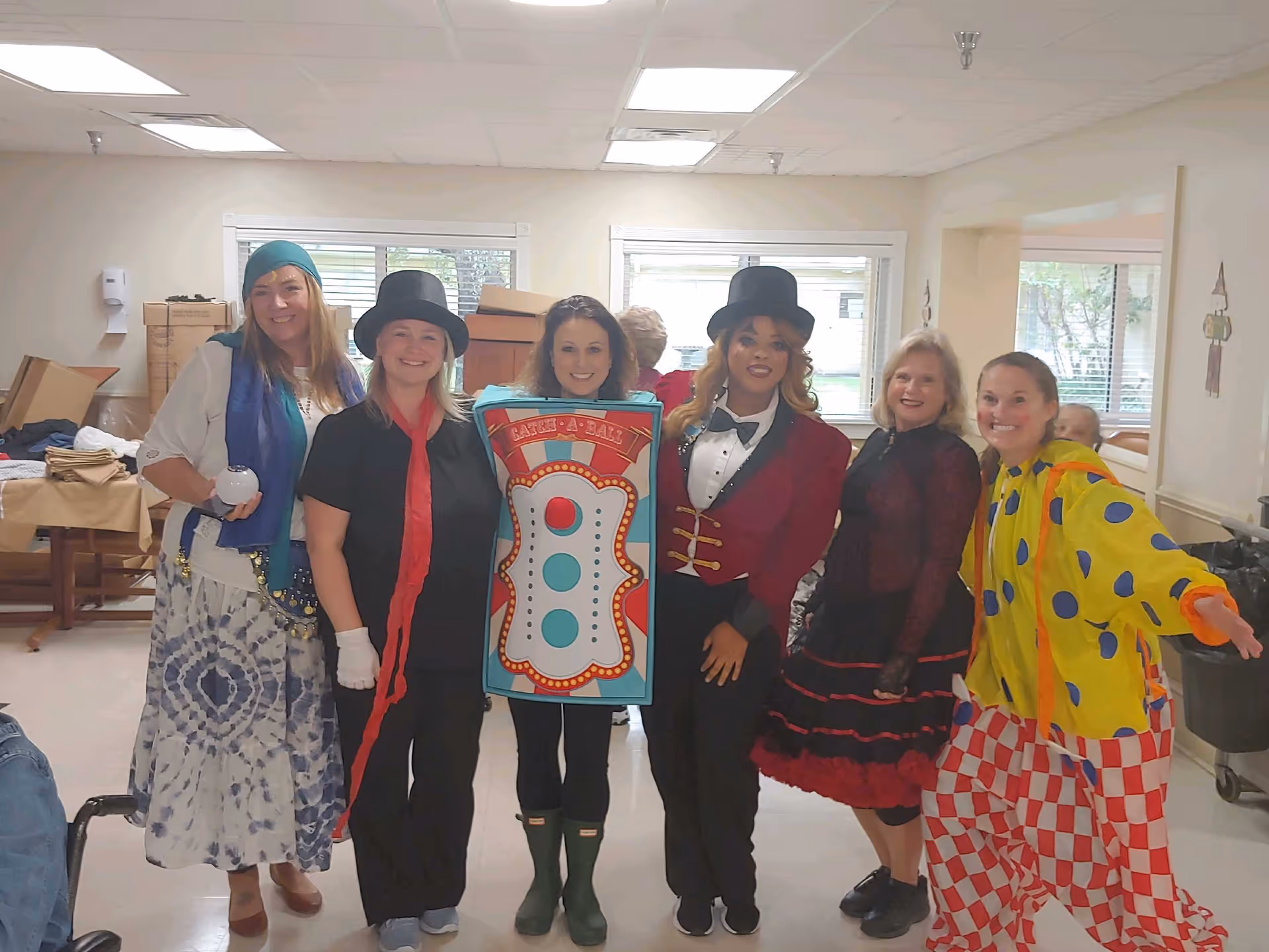 Six adults dressed in colorful costumes posing and smiling inside a room with windows and fluorescent ceiling lights. The costumes include a pirate, a person in a black outfit with a red scarf and top hat, a person dressed as a carnival game, a ringmaster, a woman in a black and red dress, and a clown in a yellow and red polka dot outfit. The room has tables and boxes in the background.