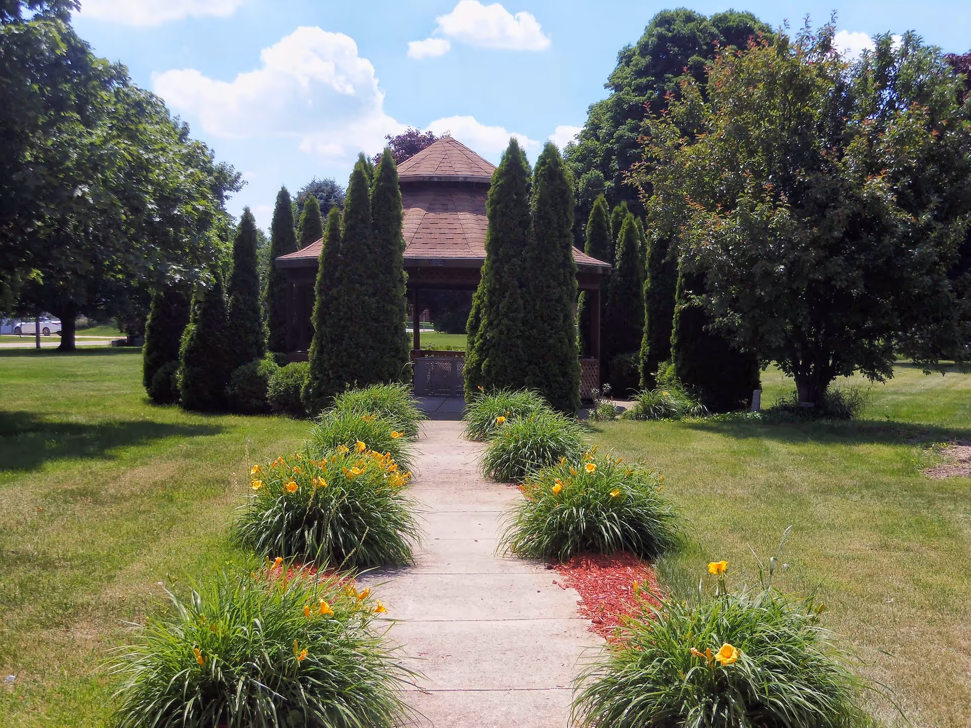 A concrete path lined with flowering shrubs leads to a wooden gazebo surrounded by tall evergreen trees in a grassy park.