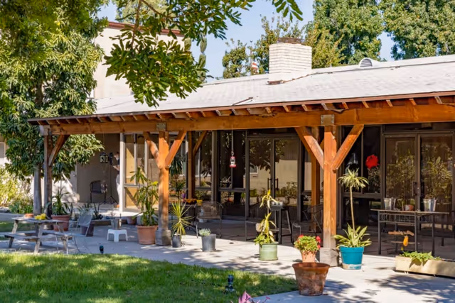 Outdoor patio area with a wooden pergola attached to a building. Several potted plants are placed on the concrete floor under the pergola and around the patio. There are glass doors and windows along the building wall, and trees provide shade in the background.