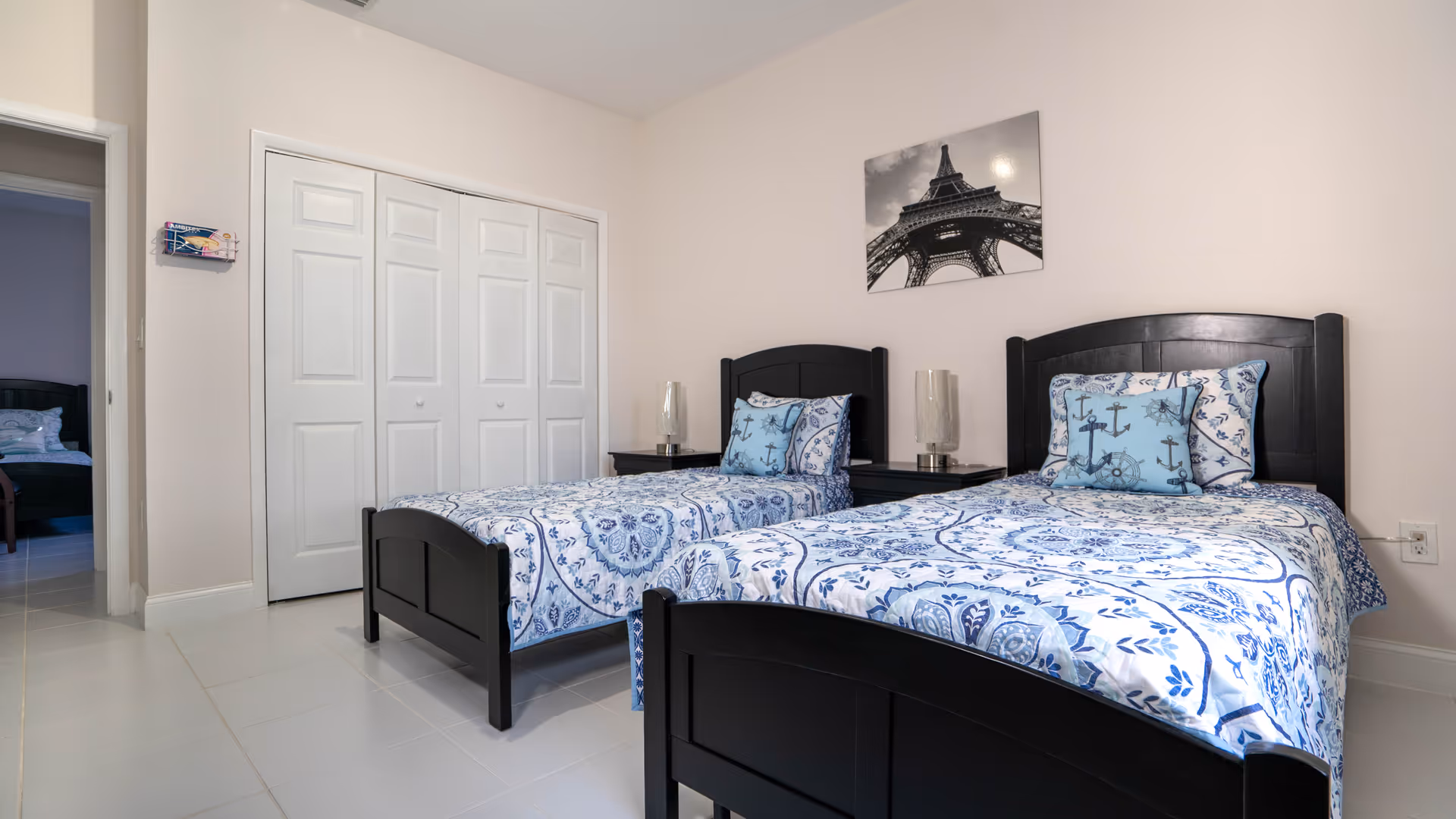 A bedroom with two twin beds featuring black wooden frames and blue patterned bedding with anchor-themed pillows. Between the beds are two black nightstands with glass lamps. A black and white photo of the Eiffel Tower hangs on the wall above the beds. There is a white closet with double doors and an open doorway leading to another room with a similar bed visible.