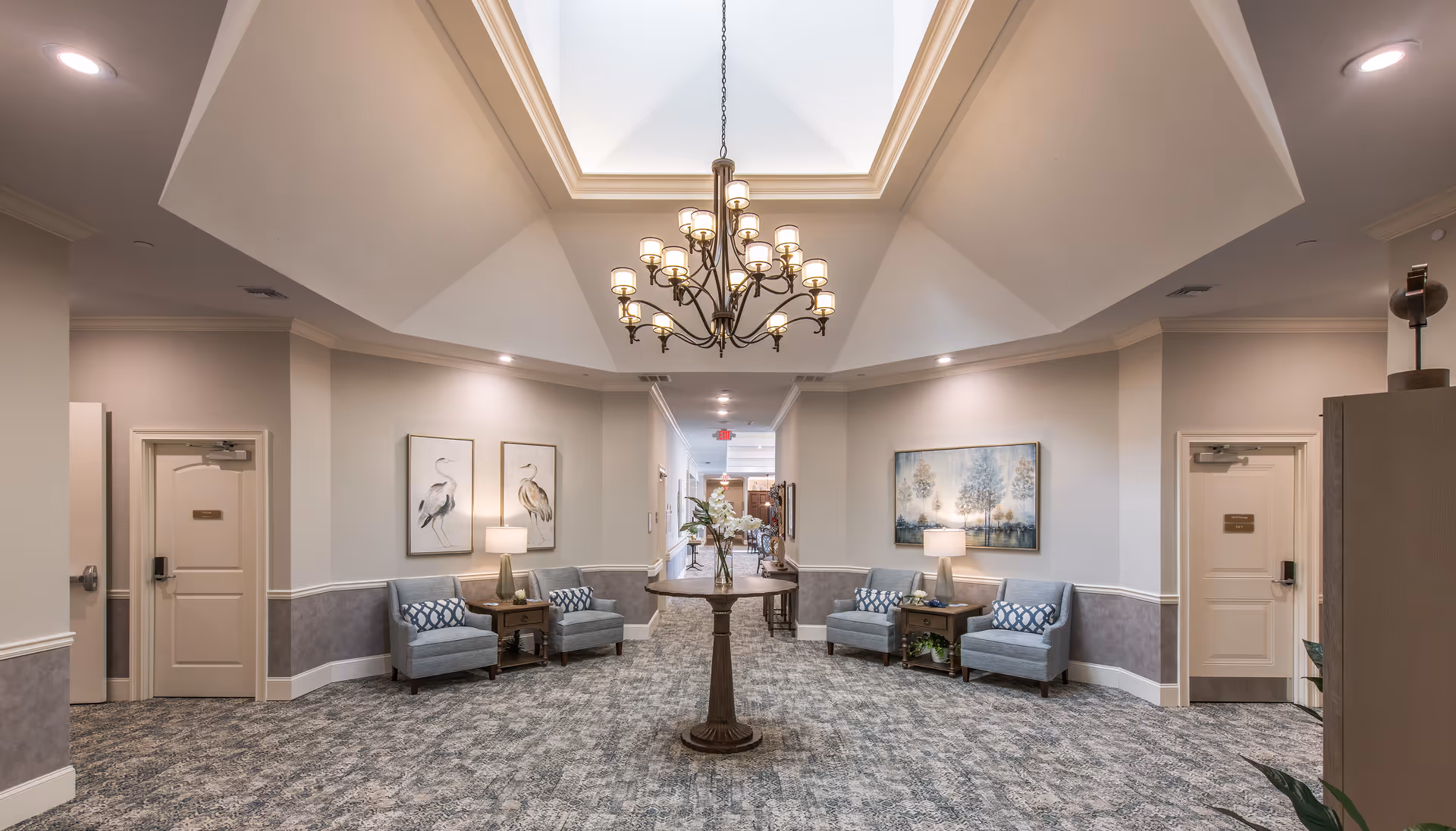 A well-lit interior hallway in a senior living facility with a central round wooden table holding a vase of flowers. The hallway features a high ceiling with a large chandelier and skylight. On either side of the hallway are seating areas with blue armchairs, side tables with lamps, and framed artwork on the walls. Doors are visible on both sides, and the carpet has a patterned design.