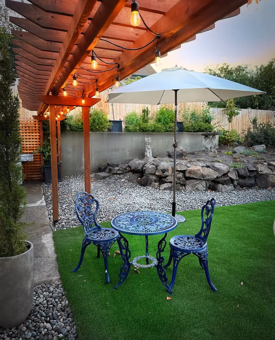Backyard patio with a blue metal table and two chairs under a white umbrella on green turf, string lights hanging from a wooden pergola and rock landscaping behind.
