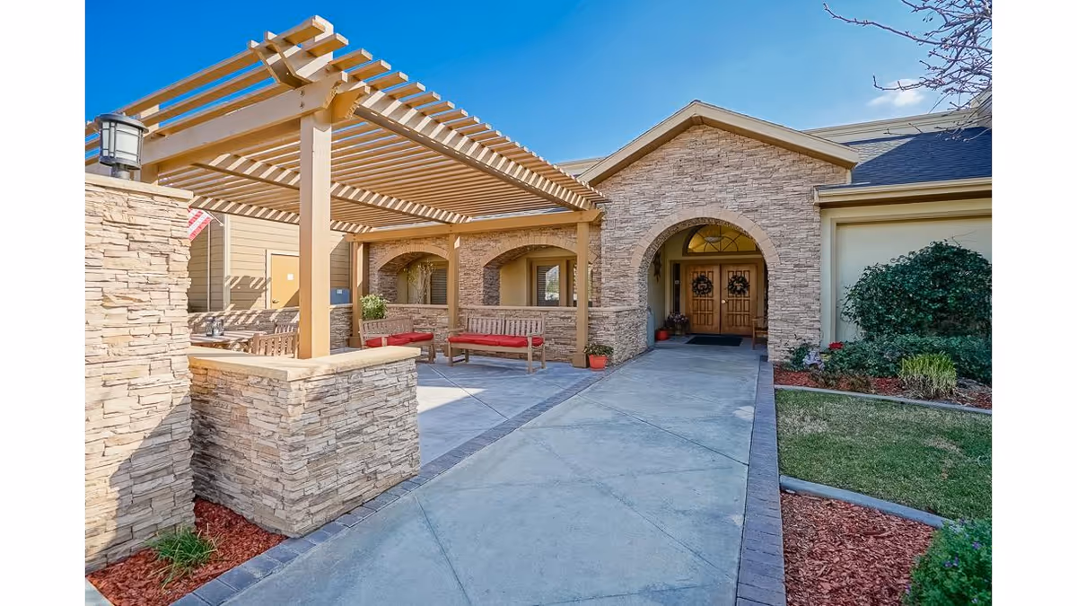 Outdoor entrance area of a senior living facility with a stone facade, a wooden pergola covering benches with red cushions, a concrete walkway leading to double wooden doors decorated with wreaths, and landscaped greenery on the side.