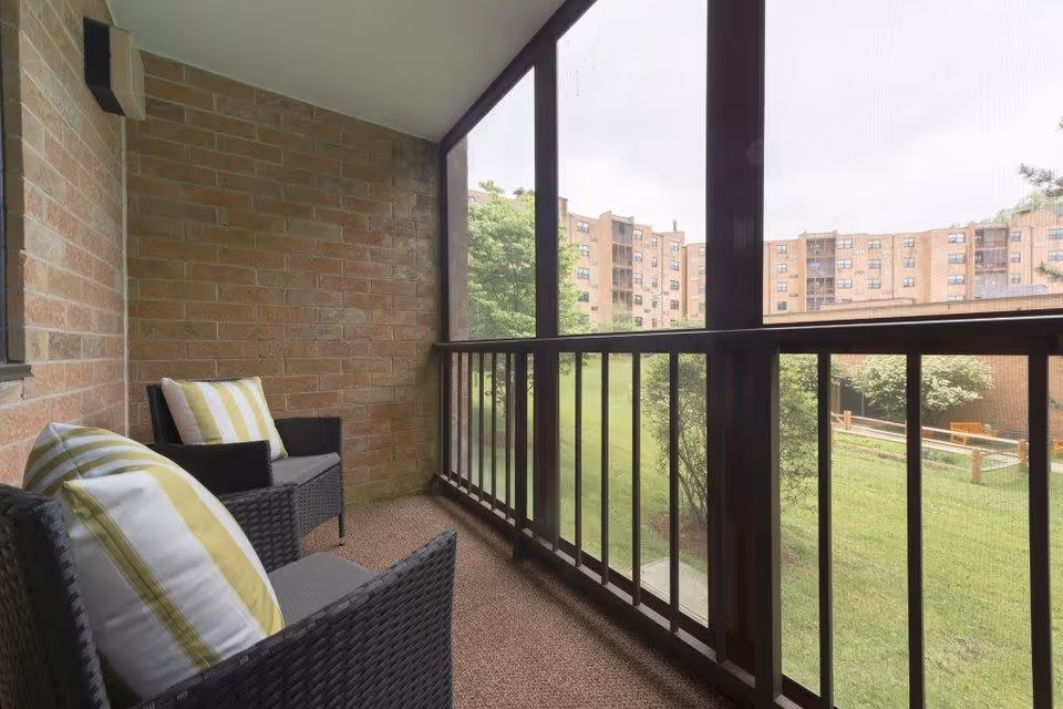 A screened-in balcony with two black wicker chairs featuring gray cushions and yellow and white striped pillows. The balcony overlooks a green lawn with trees and a multi-story brick building in the background.