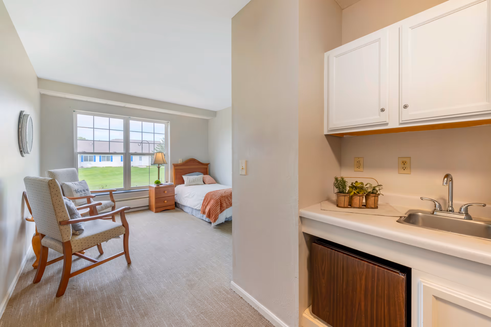 A bright senior living facility room with a single bed next to a window overlooking a green lawn. The room has two cushioned wooden chairs with pillows, a wooden nightstand with a lamp, and a small kitchenette area with a sink, countertop, and white cabinets. There are small potted plants on the countertop.