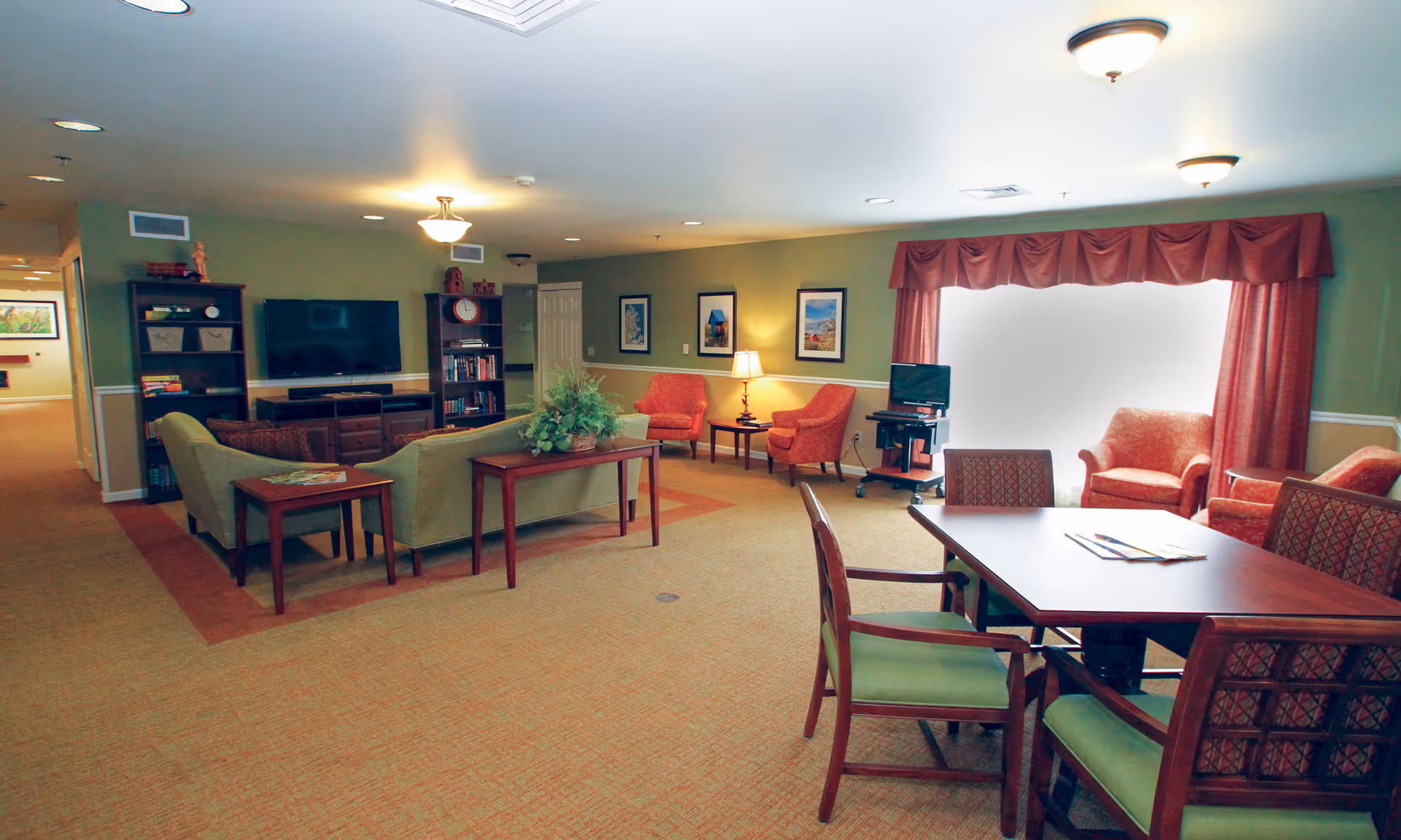 Well-lit communal living room with sofas, armchairs, a television, and a table with chairs near a large window.