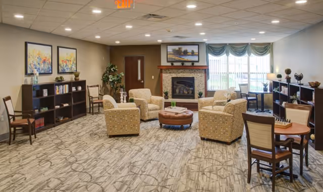 Spacious senior living common room with upholstered armchairs and an ottoman arranged around a fireplace, bookshelves, and a large window with drapes.