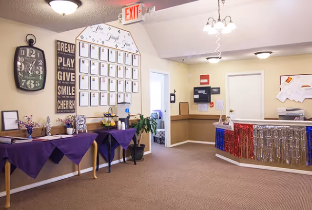 Interior view of a senior living facility reception area with a decorated front desk featuring red, white, and blue tinsel garlands. To the left, there are tables covered with purple tablecloths holding various items including flowers and a coffee station. A large clock and inspirational wall art are mounted on the wall above the tables. A bulletin board with multiple papers is visible behind the reception desk.