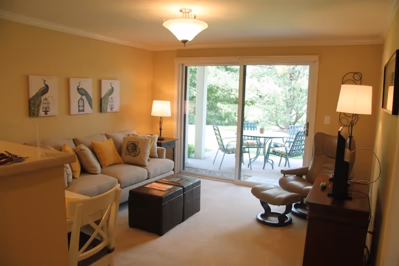A cozy living room with a beige couch adorned with yellow and patterned pillows, a matching ottoman, a leather recliner chair with an ottoman, and a wooden TV stand with a flat-screen TV. The room has warm yellow walls decorated with three peacock-themed paintings. There are two lamps providing soft lighting, and a large sliding glass door opens to an outdoor patio with a table and chairs surrounded by greenery.
