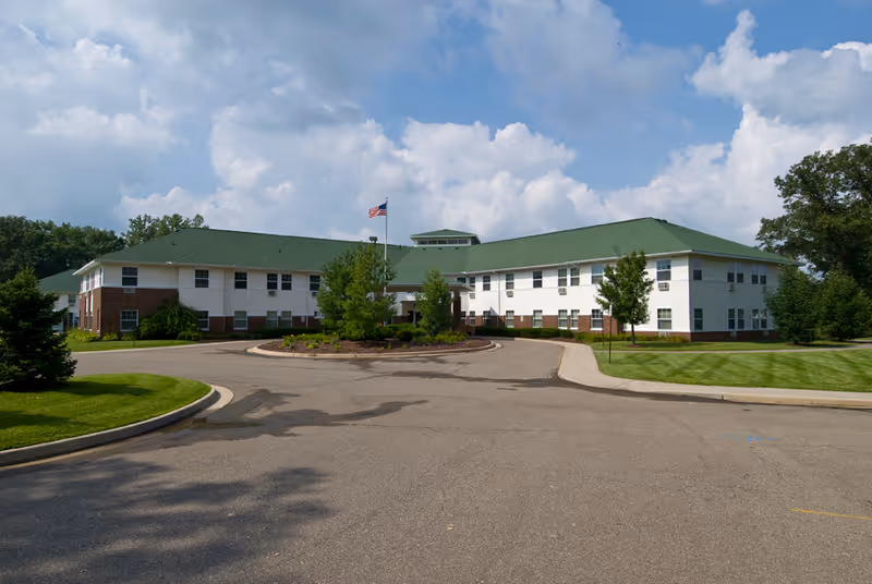 Exterior view of a two-story senior living facility building with a green roof and white and brick walls. There is a circular driveway with landscaped greenery in the center and an American flag flying on a flagpole. The sky is partly cloudy.
