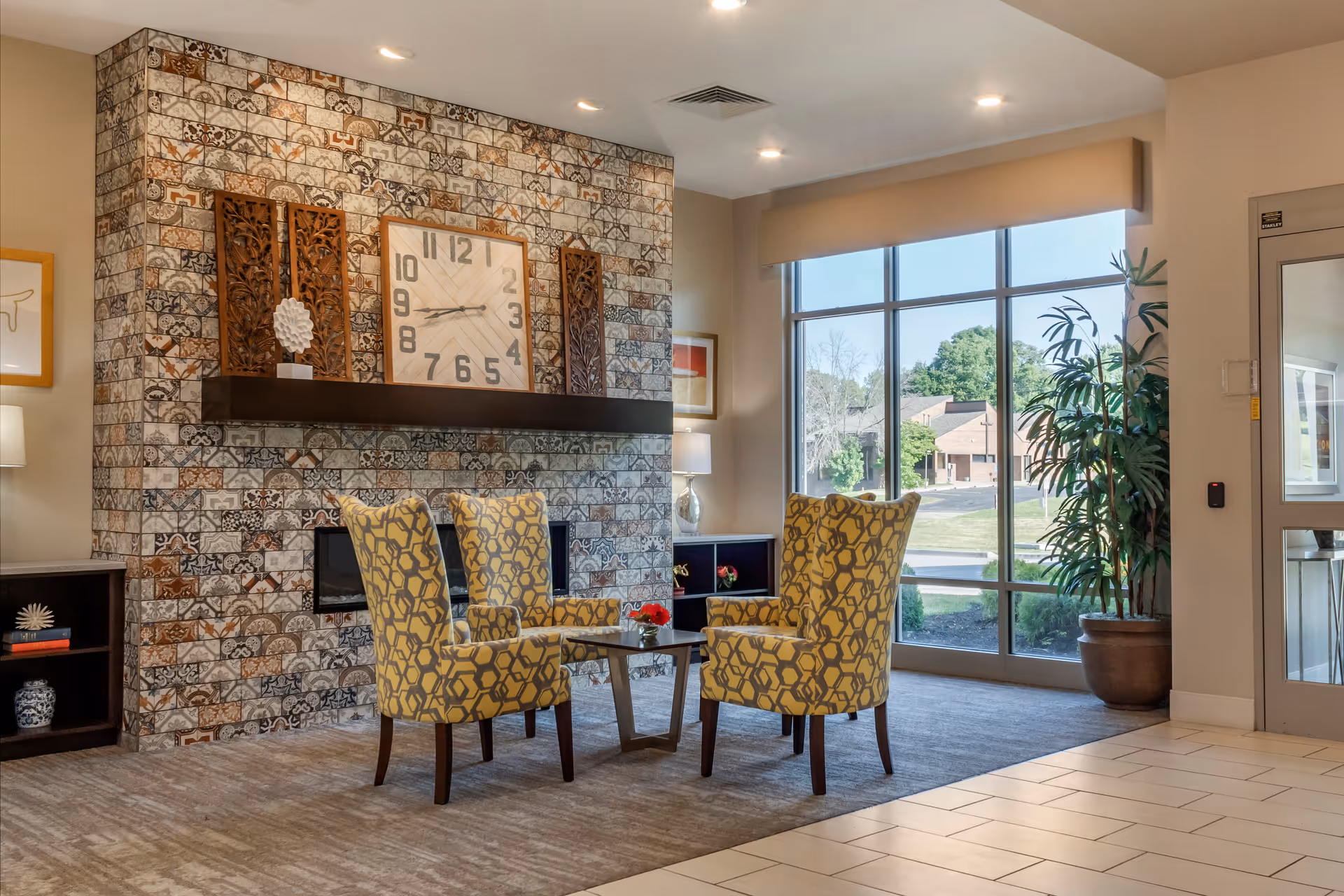 A cozy seating area in a senior living facility with four yellow patterned armchairs arranged around a small glass coffee table. Behind the chairs is a decorative brick wall with a large clock and wooden art pieces on a dark wooden mantel. To the right, large windows let in natural light and show a view of trees and buildings outside. A potted plant is near the window, and the floor is a mix of carpet and tile.