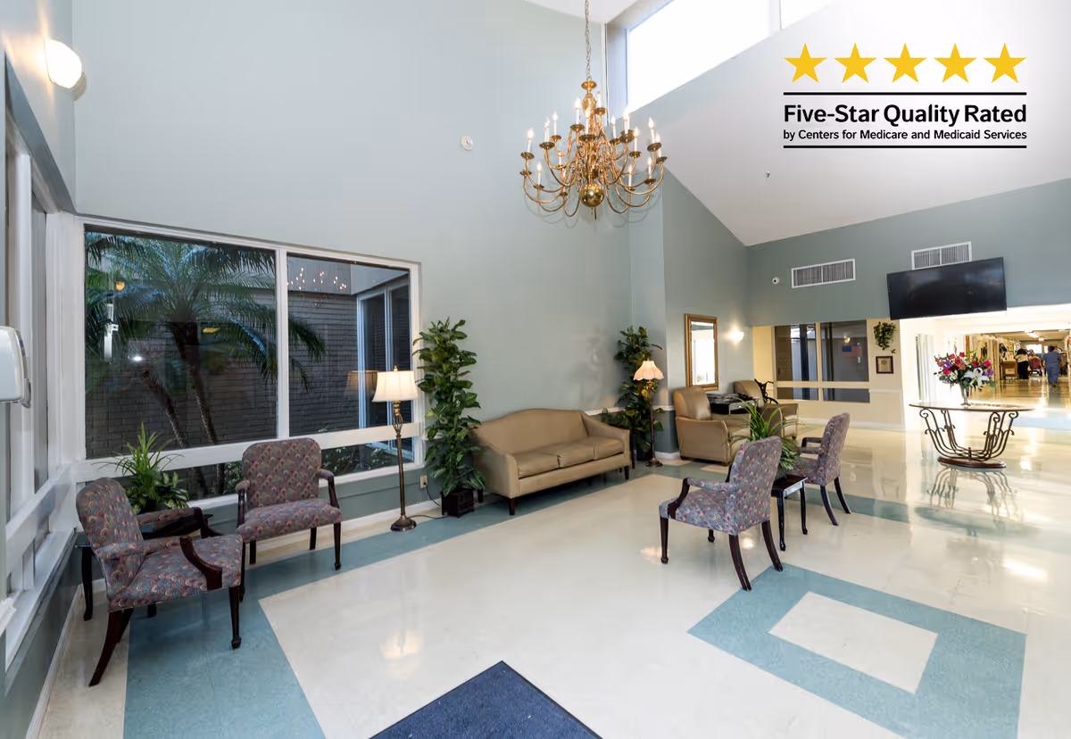A spacious and well-lit seating area in a senior living facility with several upholstered chairs and sofas arranged around the room. There are large windows showing palm trees outside, a chandelier hanging from the ceiling, potted plants, a floor lamp, and a wall-mounted TV. A table with a flower arrangement is visible in the background near a hallway.