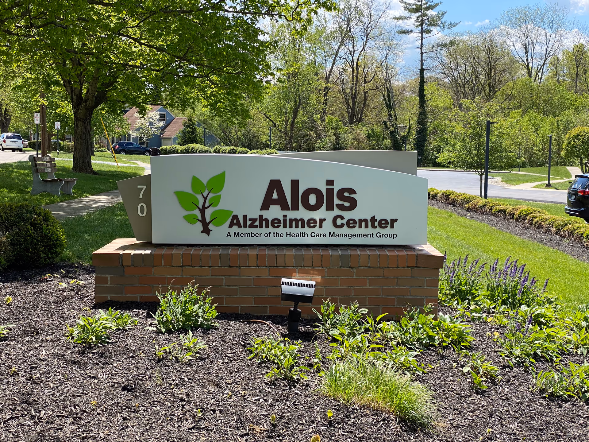 A landscaped brick entrance sign reading "Alois Alzheimer Center" with a leaf logo beside a tree-lined road.