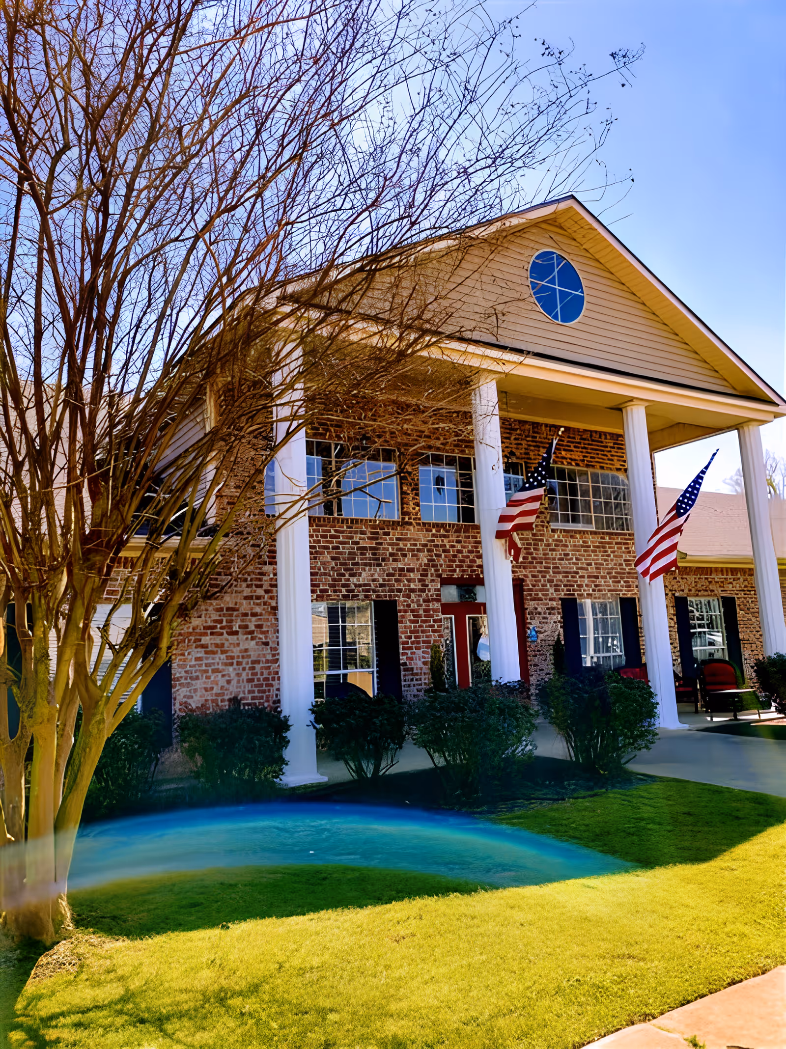 Front exterior of a two-story brick building with tall white columns, American flags, and a manicured lawn.
