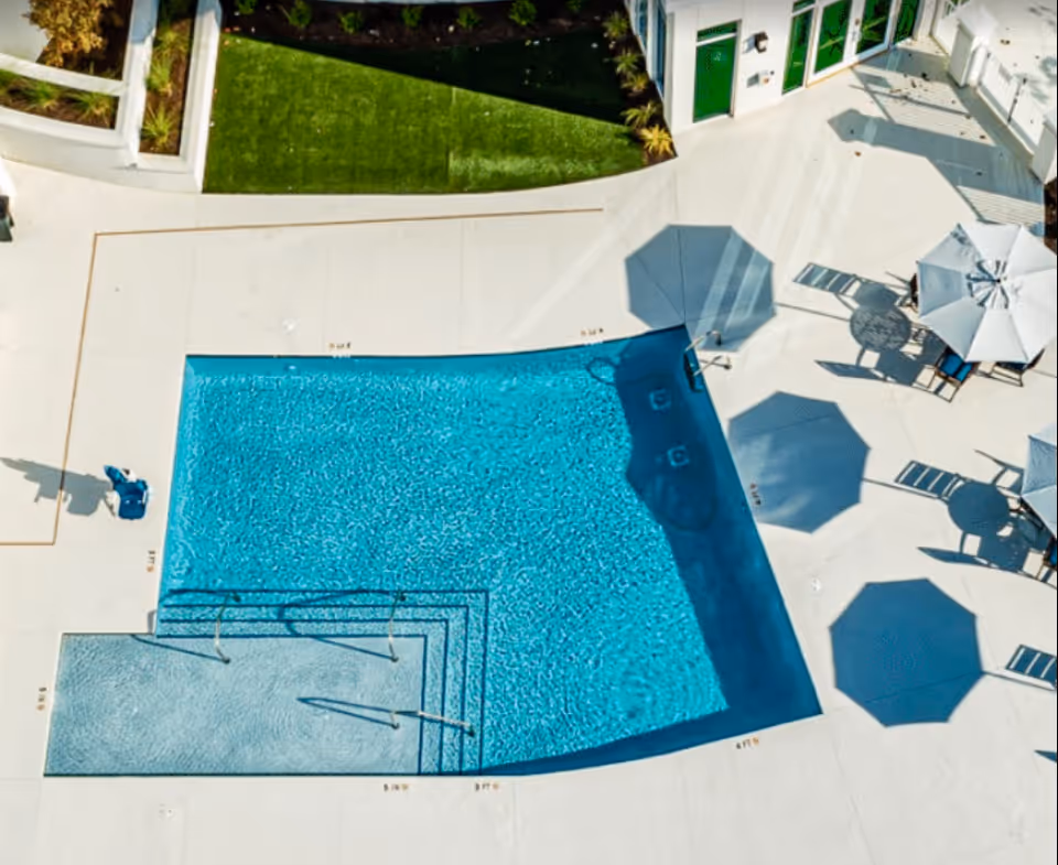 Aerial view of a rectangular outdoor swimming pool with clear blue water, surrounded by a white concrete deck. There are several white umbrellas casting shadows on the deck, along with tables and chairs arranged nearby. A patch of green grass and some plants are visible at the top edge of the image.