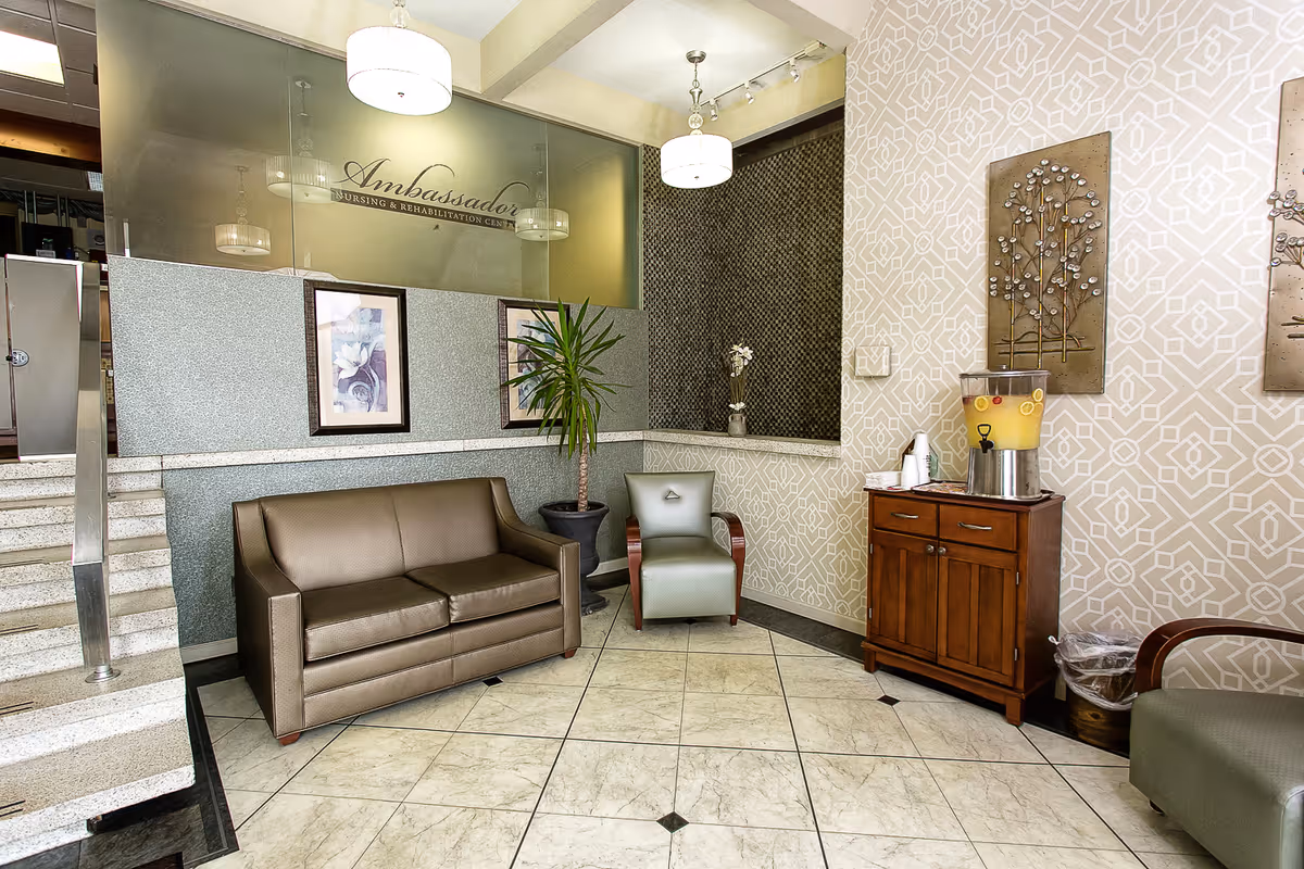 Lobby seating area with a leather sofa and chairs, a potted plant, decorative wall art and a wooden cabinet with a beverage dispenser beneath glass signage that reads 'Ambassador'.